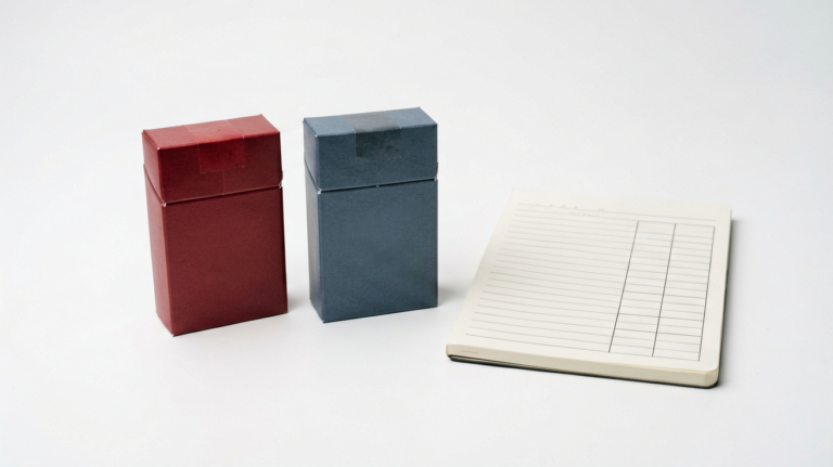 Unbranded red and blue cigarette cartons beside a price comparison notebook